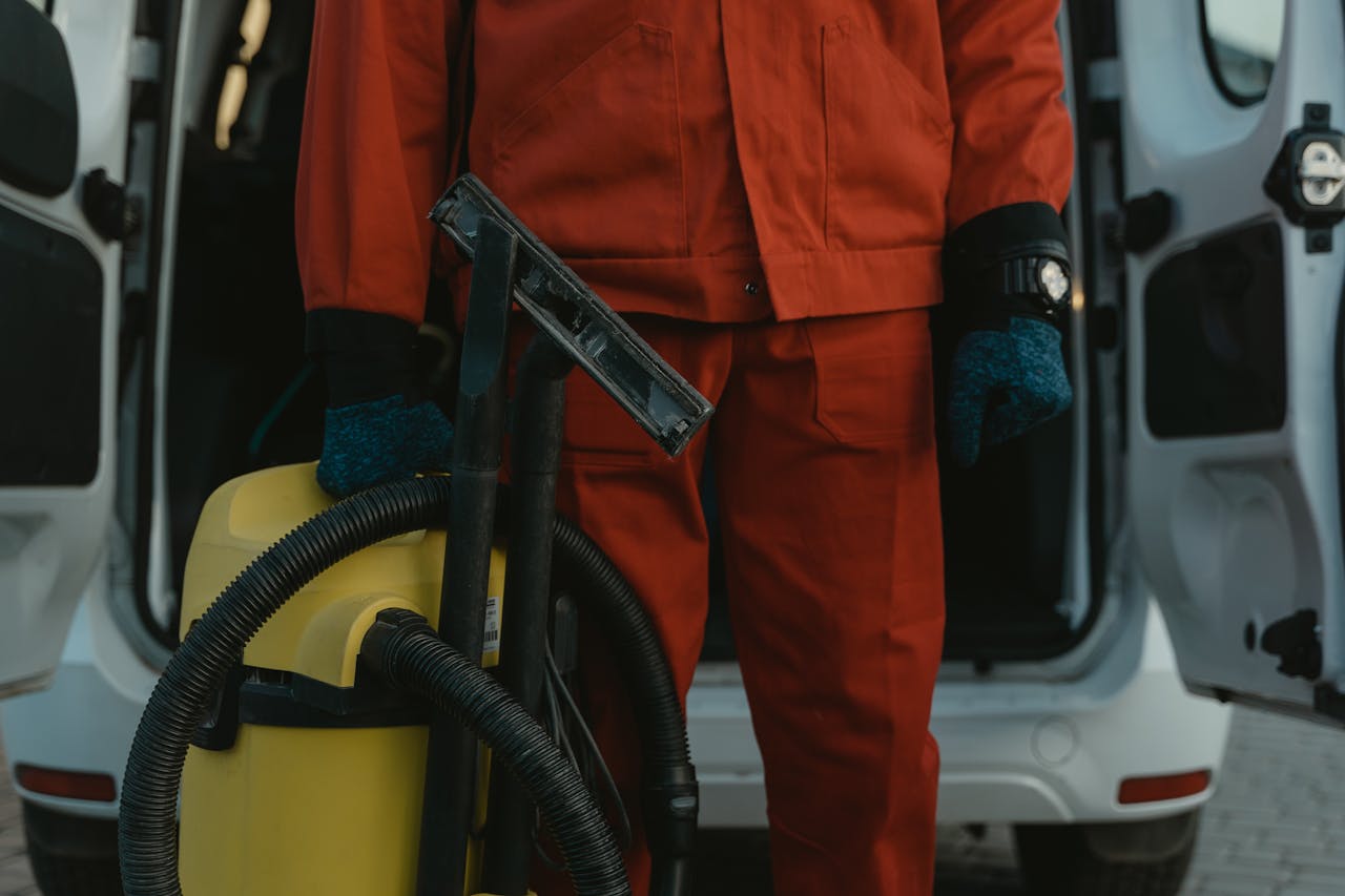 A professional cleaner in a red uniform holding a vacuum near a van, ready for service.